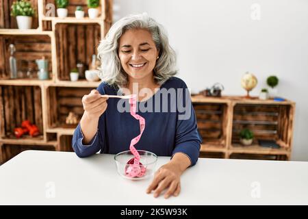 Femme d'âge moyen à cheveux gris mangeant un mètre-ruban assis sur la table à la maison. Banque D'Images