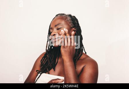 Femme mature avec des dreadlocks appliquant de la crème hydratante sur son visage. Femme à la peau sombre confiante prenant soin de sa peau mélanée sans défaut. Maturité Banque D'Images
