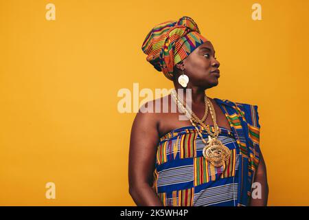 Femme africaine élégante portant une tenue traditionnelle sur fond jaune. Femme noire mûre vêtue de tissu Kente coloré et de bijoux dorés Banque D'Images