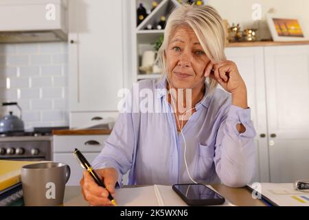 Femme caucasienne âgée portant des écouteurs, assise à une table dans la cuisine, ayant un appel vidéo Banque D'Images