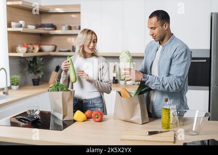 Portrait d'un jeune couple heureux et marié qui rentre à la maison après avoir fait du shopping et déballé des sacs en papier avec des provisions dans la cuisine. Concept de ménage et de relation. Banque D'Images