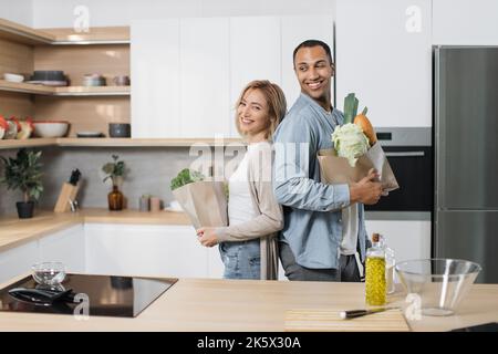 Positif joyeux jeune indien et blonde femme debout ensemble dans la cuisine, souriant, se tenant dos à dos et déballant le sac de papier d'épicerie. Concept de famille, de modes de vie domestiques et de saine alimentation Banque D'Images