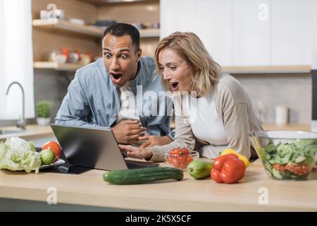Belle femme indienne excitée et blonde caucasienne joyeuse ayant de bonnes nouvelles en utilisant des recettes de recherche d'ordinateur portable, en commandant des achats en ligne, en regardant les cours de cuisine pendant la préparation de délicieux salade végétalienne Banque D'Images