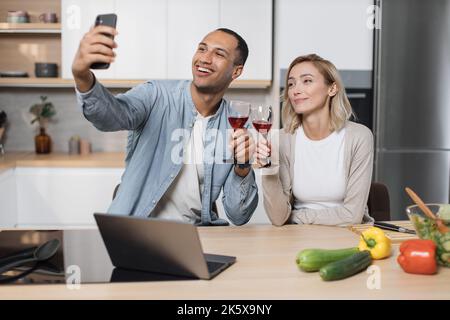 Mignon couple multinational prenant le selfie ou d'avoir la conversation vidéo avec des amis tout en ayant un petit déjeuner sain à la maison, en utilisant le tout nouveau téléphone mobile, intérieur de cuisine, espace de copie Banque D'Images