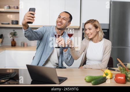 Mignon couple multinational prenant le selfie ou d'avoir la conversation vidéo avec des amis tout en ayant un petit déjeuner sain à la maison, en utilisant le tout nouveau téléphone mobile, intérieur de cuisine, espace de copie Banque D'Images