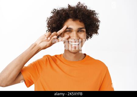 Portrait en gros plan d'un homme hispanique souriant montrant la paix, le V-sign près des yeux et regardant heureux, les gens positifs et les émotions, debout sur blanc Banque D'Images