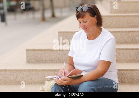 La femme mature travaille à distance, l'enseignement supérieur pour les adultes, la femme décontractée écrit et note dans un carnet assis sur les escaliers à l'extérieur, freelance.C Banque D'Images