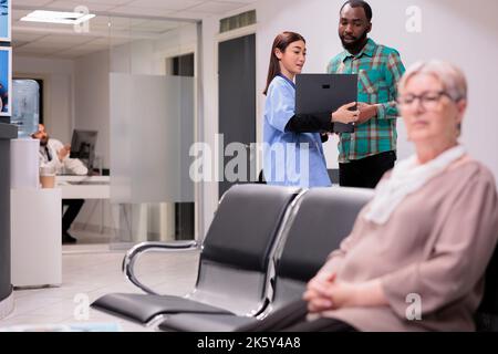 Clinique médecin du service médical d'urgence en charge de la recherche de jeunes afro-américains de dossiers électroniques sur ordinateur portable. Femme caucasienne âgée assise en attente d'admission à l'hôpital. Banque D'Images