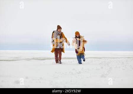 Vue de face portrait d'un jeune couple insouciant qui court vers l'extérieur en hiver au bord d'un lac gelé, espace de copie Banque D'Images