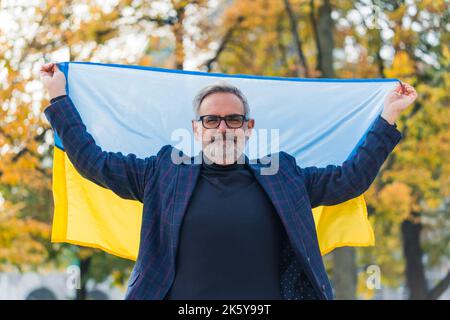Portrait d'extérieur sain de l'homme barbu caucasien et touché dans un blazer sombre regardant la caméra pleine de larmes et tenant le drapeau ukrainien bleu-et-jaune avec les deux mains. Photo de haute qualité Banque D'Images
