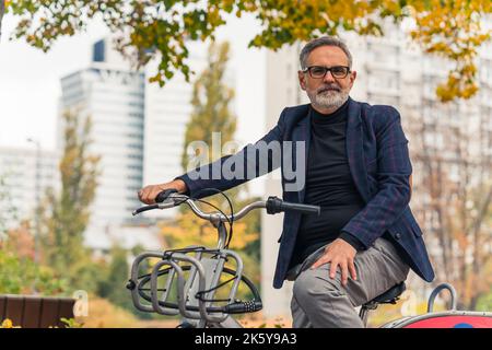 Mi-plein dans le parc. Satisfait heureux barbu gris-cheveux homme caucasien dans son 60s regardant la caméra tout en étant assis sur le vélo gris. Bâtiments flous et arbres d'automne en arrière-plan. Photo de haute qualité Banque D'Images