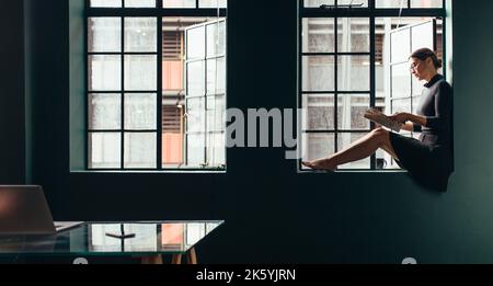 Vue latérale d'une femme assise par la fenêtre et lisant un magazine. Jeune femme asiatique avec magazine dans son bureau. Banque D'Images