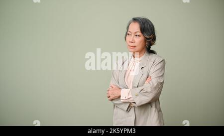 Insatisfait, malheureux et contrarié 60s ans femme asiatique ou femme d'affaires, regardant loin de l'appareil photo, bras pliés, debout isolé sur fond vert. Banque D'Images