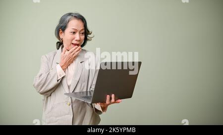 Stupéfait et surpris 60s femme ou femme d'affaires asiatique âgée tenant et regardant l'écran d'ordinateur portable, se tenant isolé sur fond vert. Choqué, sauf Banque D'Images