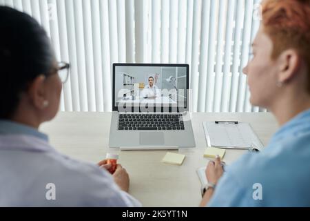 Professionnels de la santé assis à une table devant l'ordinateur portable et parlant en ligne à un collègue pendant un appel vidéo Banque D'Images