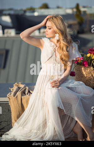 Belle femme en robe blanche élégante s'asseoir sur une couverture et avec le regard tourné de la tête au coucher du soleil sur la terrasse de toit. Une femme élégante se détend avec un verre de champagne Banque D'Images