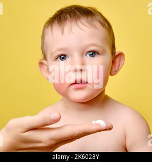 La mère sent la crème allergique sur le visage du bébé tout-petit, fond jaune studio. Portrait en gros plan d'un bébé mignon et d'une femme main avec de la crème enfant âgé d'un an Banque D'Images