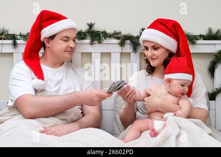 Le mari donne de l'argent à la femme avec l'enfant sur le lit décoré pour noël et le nouvel an, couple de famille dans la chambre à la maison Banque D'Images