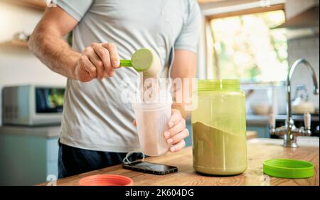 Gros plan d'un homme caucasien versant une boule de poudre de protéine de lactosérum de chocolat à un shake de santé pour l'énergie pour l'entraînement d'entraînement dans une cuisine à la maison Banque D'Images