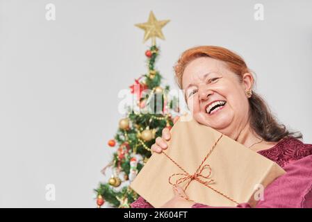 Bonne femme hispanique mature tenant le cadeau de Noël qu'elle vient de recevoir, souriant largement. La joie des vacances. Banque D'Images