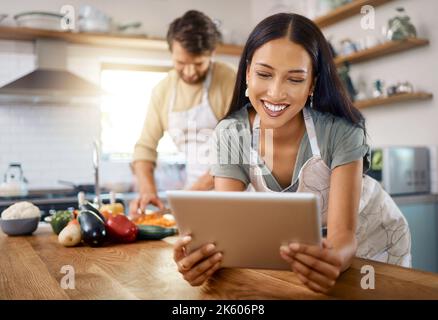 Gros plan sur une jeune femme heureuse qui navigue sur une tablette numérique tandis que son petit ami cuisine en arrière-plan. Jeune femme hispanique à la recherche de recette à Banque D'Images