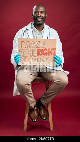 Un médecin afro-américain covid tient et montre une affiche. Portrait complet d'un médecin noir souriant isolé sur fond rouge de studio avec Banque D'Images