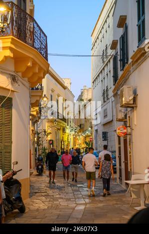 Touristes et habitants marchant le long d'une rue typique étroite et pavée dans la vieille ville de Gallipoli en début de soirée, Apulia (Puglia), Italie. Banque D'Images