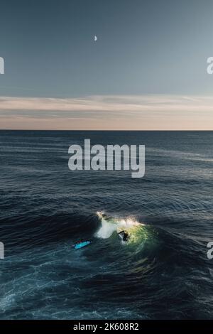 Surfeur aventureux attrapant une vague sous le ciel du soir, avec une toile de fond de lune et de falaises lointaines, capturant l'essence du surf, en Cornouailles. Banque D'Images