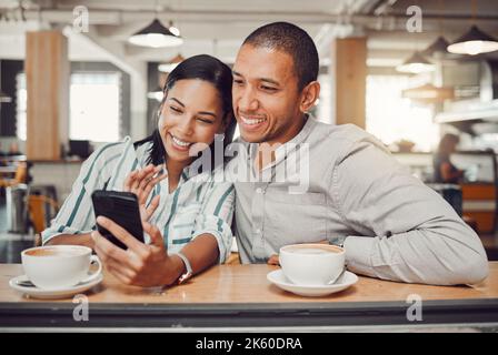 Joyeux jeune couple mixte de course assis à table prenant un café tout en regardant quelque chose sur smartphone dans le café. Couple aimant souriant en prenant Banque D'Images
