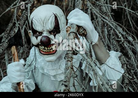 gros plan d'un clown maléfique créepy dans les bois au crépuscule, portant un costume gris avec une ruff blanche, avec une hache dans la main à l'observateur Banque D'Images