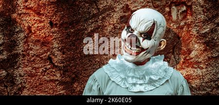 un clown maléfique, portant un costume gris avec une ruff blanche, se pose à l'observateur avec un sourire effrayant, devant le mur rouillé d'une maison abandonnée, Banque D'Images