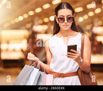 Jeune femme élégante portant des lunettes de soleil à l'aide d'un téléphone portable tout en portant des sacs pendant une frénésie de shopping. Jeune femme brunette envoyant un texte avec elle Banque D'Images