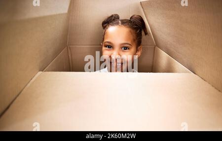 Portrait d'une petite fille hispanique mignonne jouant avec une boîte en carton dans un nouvel appartement. Jolie fille de race mixte se cachant dans une boîte et souriant dans une maison Banque D'Images