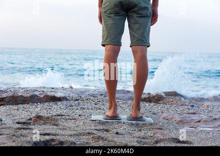 Un jeune homme fait du yoga en se tenant debout sur les ongles. Un yogi se tient sur une planche avec des ongles sur une plage de mer. Yoga, méditation sur fond de vague de mer Banque D'Images