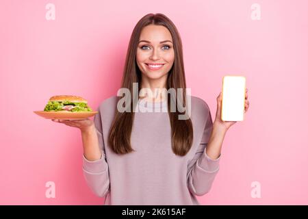 Photo d'une jolie personne positive main tenir la plaque de hamburger montrant l'espace vide téléphone écran isolé sur fond de couleur rose Banque D'Images