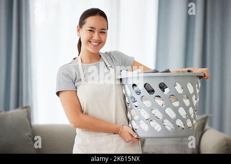 Portrait d'une belle jeune femme de race mixte tenant un panier à linge tout en nettoyant son appartement. Une femme asiatique heureuse souriant en pliant Banque D'Images