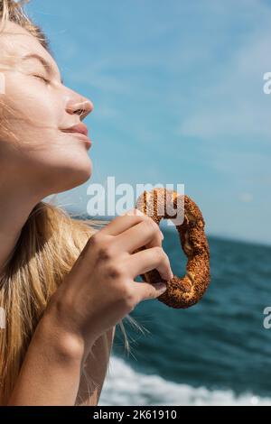 femme blonde heureuse avec des yeux fermés tenant bagel de sésame turc près de la mer, image de stock Banque D'Images