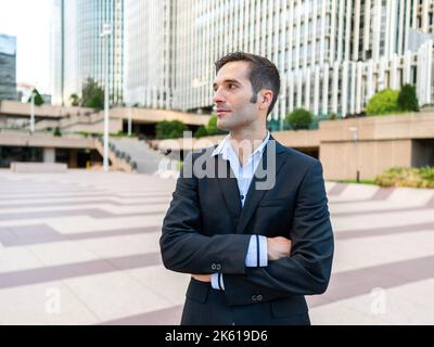 Homme adulte élégant et confiant, en costume formel, debout près du bâtiment moderne de la rue de la ville et donnant sur l'extérieur Banque D'Images