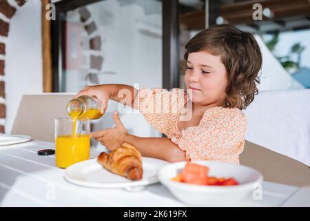 Dorer l'enfant aux cheveux assis à table et verser le jus d'orange dans le verre pendant le petit-déjeuner avec croissant et salade de fruits au café Banque D'Images