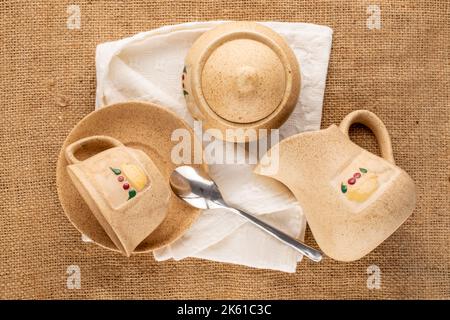 Une tasse d'argile sur soucoupe, sucrier et pot à lait avec serviette en lin blanc sur toile de jute, macro, vue du dessus. Banque D'Images