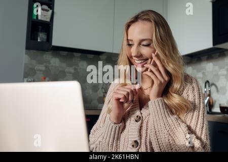 Rencontres en ligne. Jolie jeune femme parle au téléphone tout en regardant son écran d'ordinateur portable, assis dans sa cuisine. Banque D'Images