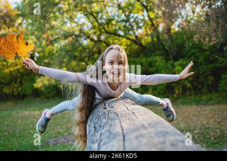 Une jolie fille se trouve sur une bûche tombée dans le parc avec ses bras écartés comme des ailes. Fille en équilibre sur une bûche avec une feuille dans sa main. Banque D'Images