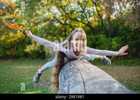 Une jolie fille se trouve sur une bûche tombée dans le parc avec ses bras écartés comme des ailes. Fille en équilibre sur une bûche avec une feuille dans sa main. Banque D'Images