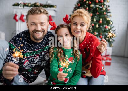 Joyeux famille dans les sweaters de noël tenant des sparkers et regardant l'appareil photo à la maison, image de stock Banque D'Images