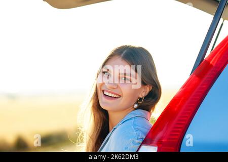 Bonne jeune femme regardant la caméra près de la voiture au coucher du soleil doré dans le champ Banque D'Images