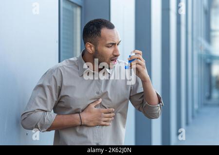 Un homme à l'extérieur d'un immeuble de bureaux a une grave crise d'asthme, un homme d'affaires a de la difficulté à respirer, un travailleur dans une chemise utilise un inhalateur pour faciliter la respiration, un homme hispanique dans une chemise décontractée. Banque D'Images