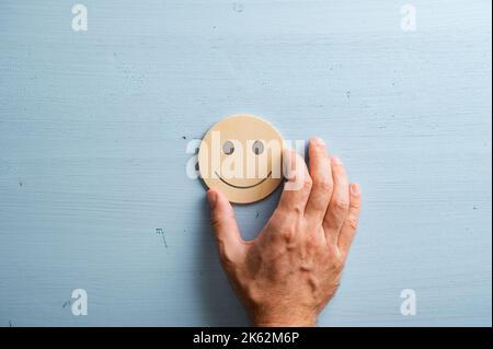 Main masculine plaçant un visage souriant coupé dans un cercle de bois coupé sur fond bleu pastel. Banque D'Images