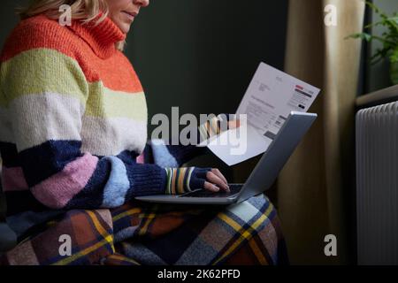 Femme en gants avec ordinateur portable et Bill essayant de rester au chaud par le radiateur pendant la crise énergétique du coût de la vie Banque D'Images