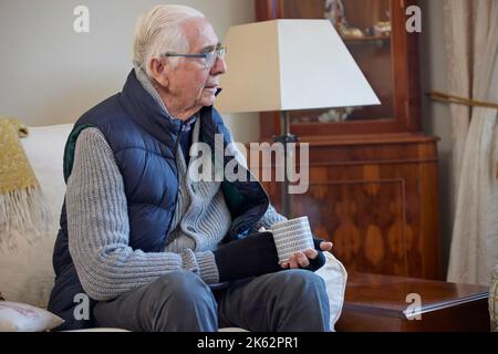 Homme senior portant des vêtements supplémentaires avec boisson chaude essayant de rester au chaud à la maison en crise d'énergie Banque D'Images