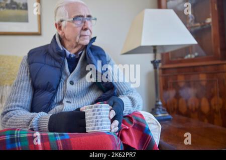 Homme senior portant des vêtements supplémentaires avec boisson chaude essayant de rester au chaud à la maison en crise d'énergie Banque D'Images